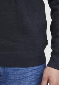 Mörkgrå stickad tröja med ribbade muddar och nederkant, tillsammans med blå denimjeans. Har en liten svart etikett på sidan.