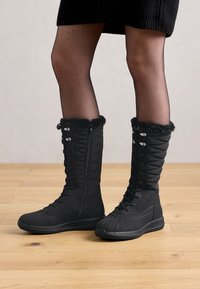 Botas negras hasta la rodilla con paneles acolchados, un suave ribete de piel en la parte superior y un frente con cordones, con una suela de goma plana para mayor agarre.