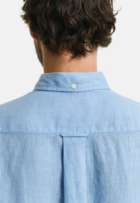 Camisa de lino azul claro con cuello abotonado, detalle de pliegue en la parte trasera y un cierre de botón en el cuello.