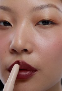 Gros plan d'une personne appliquant un rouge à lèvres rouge foncé avec un applicateur beige sur ses lèvres.