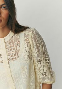 Mežģīņu pogājams tops maigi dzeltenā krāsā ar ziedu rakstiem, caurspīdīgu tekstūru un uzpūstām piedurknēm, ar apaļu apkakli un smalkām detaļām.