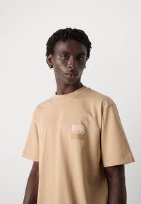 Bežinis t-shirt, pagamintas iš medvilnės, su apvaliu kaklu, laisvu kirpimu ir mažos šviesesnės spalvos logotipo etiketėmis kairėje krūtinės pusėje.