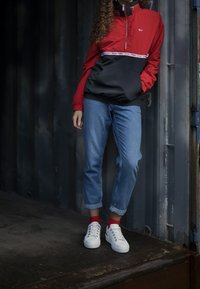 Osoba oblečená v červené a černé bundě Tommy Jeans, modrých roztržených džínách, červených ponožkách a bílých teniskách, stojí proti tmavé kovové zdi.