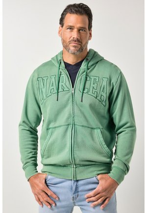Mann mit kurzen dunklen Haaren und Bart, trägt einen grünen Kapuzenpullover mit Reißverschluss und dem Schriftzug "NARCEA" sowie hellblaue Jeans, steht mit den Händen nahe den Taschen.