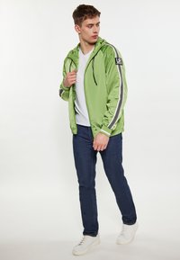 Grön nylonjacka med vita och svarta randiga detaljer, justerbar huva, dragkedjestängning, bärs över en vit t-shirt och mörka jeans.