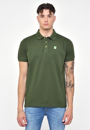 Polo verde con colletto, tre bottoni e un piccolo logo sul petto. Realizzato in tessuto liscio. Indossato con jeans azzurri chiari.