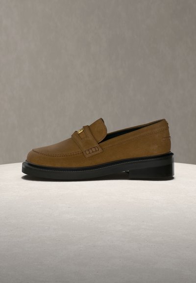 Brun ruskindslipper med sort sål og guldfarvet spænde på toppen, vist mod en neutral grå og hvid baggrund.