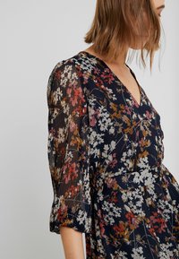 Blommig klänning med djup V-ringning, med korta, plisserade, genomskinliga ärmar. Mörk marin bakgrund med mångfärgade blommönster.