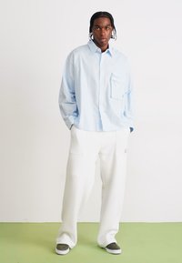 Lysblå oversized skjorte med en brystlomme, parret med hvide breddebenede bukser med forlommer. Grå sneakers fuldender outfittet.