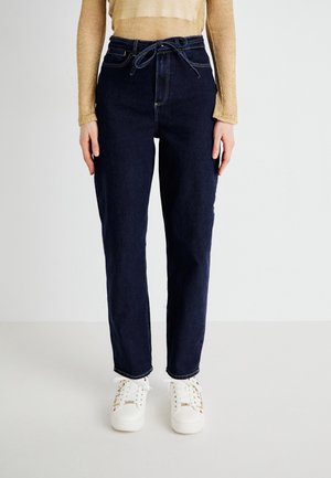 Vrouw draagt donkerblauwe high-waisted jeans met een trekkoord in de taille, een beige top met lange mouwen en witte sneakers met patroonveters.