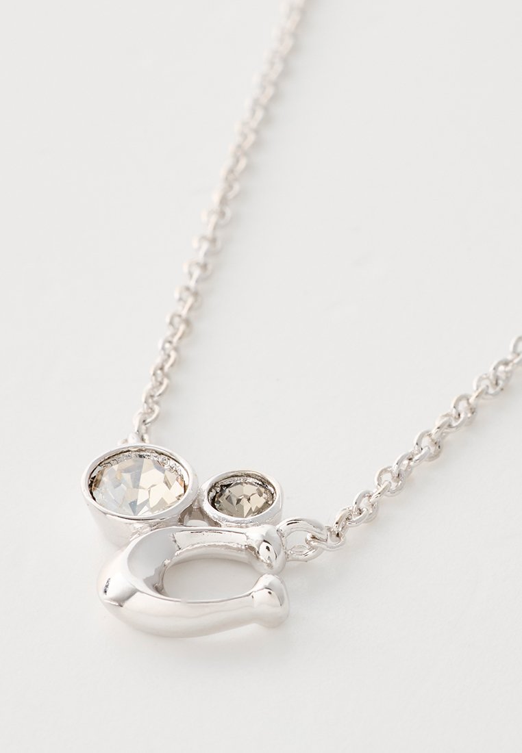 Collier en argent avec un design entrelacé, comportant deux gemmes transparentes—une grande, une petite—serties dans un métal lisse. La chaîne a un style de maillon délicat.