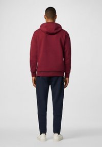 Sudadera burdeos con una capucha grande, puños y dobladillo acanalados. Combinada con pantalones azul marino y zapatillas blancas. Textura suave, diseño minimalista.