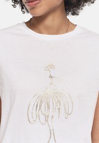 T-shirt blanc en coton doux avec une illustration de robe métallique dorée au centre. Encolure ronde, manches courtes, texture lisse.