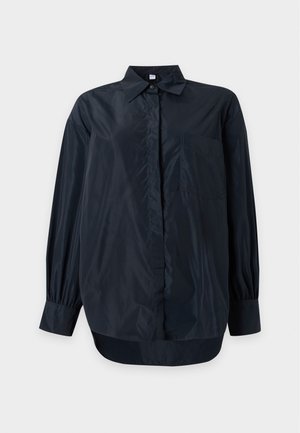 Personne portant une chemise noire ample boutonnée, un jean bleu clair à jambes larges et des baskets foncées, debout devant un fond blanc uni.