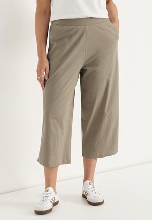 Osoba nosząca oliwkowo-zielone szerokie spodnie 7/8, białe trampki z brązowymi paskami i biały top, stojąca na jednolitym tle.