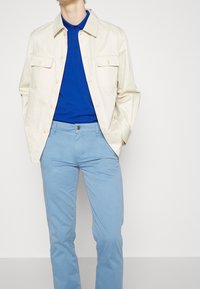 Ljus beige jacka med två bröstfickor över en blå polo-tröja, ihop med ljusblå slim-fit byxor. Slät tyg, avslappnad design.