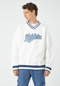 Sudadera blanca con franjas azules y el texto "Athletic" en el pecho. Tejido suave, corte holgado, puños y dobladillo acanalados. Combinada con pantalones vaqueros azules.