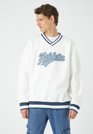 Witte sweatshirt met blauwe strepen en de tekst "Athletic" over de borst. Zacht materiaal, losse pasvorm, ribgebreide manchetten en onderrand. Draagt met een blauwe spijkerbroek.