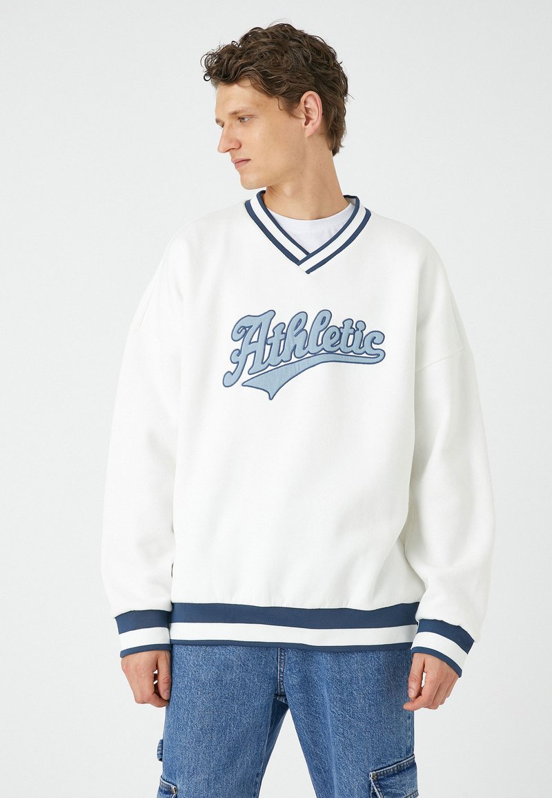 Sudadera blanca con franjas azules y el texto "Athletic" en el pecho. Tejido suave, corte holgado, puños y dobladillo acanalados. Combinada con pantalones vaqueros azules.