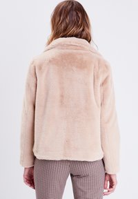 Veste en fausse fourrure beige clair avec une texture douce, un col et une coupe décontractée. La vue arrière montre les coutures et de légères variations de couleur.