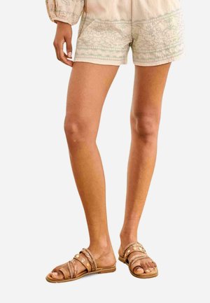 Piernas femeninas con pantalones cortos bordados beige y sandalias planas marrones con tiras trenzadas y con tachuelas sobre fondo blanco.