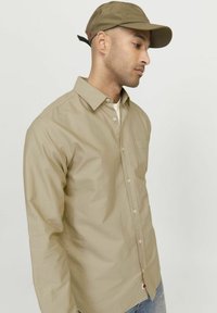 Khaki srajca z dolgimi rokavi iz gladkega materiala z ovratnikom, ki se zapenja z gumbi, in žepom na prsih. Ujemajoča se khaki kapa z rdečim poudarjanjem.
