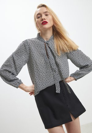Blusa a fantasia geometrica in bianco e nero con scollo a cravatta, maniche a palloncino, e gonna nera con bottoni frontali.