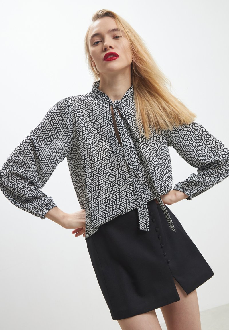 Blusa a fantasia geometrica in bianco e nero con scollo a cravatta, maniche a palloncino, e gonna nera con bottoni frontali.