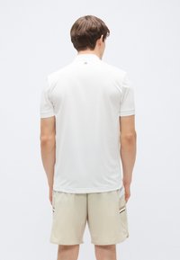 Άσπρο πόλο με κοντό μανίκι και ομοιόμορφη υφή, με μικρό λογότυπο στο πίσω μέρος. Φοριέται με μπεζ σορτς που έχει μαύρες και άσπρες λωρίδες.