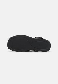 Svart sandal med flat gummisåle, justerbar stropp og glatt tekstur. Har et minimalistisk design og subtil merkevare på sålen.