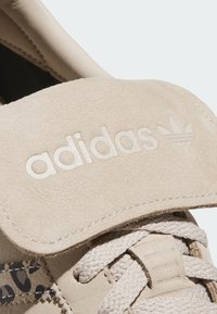 Bež antilop tenisice s podstavljenim jezikom. Sadrži urezani logo "adidas" i uzorkovane akcentne detalje sa strane. Tekstuirane petlje za vezice.