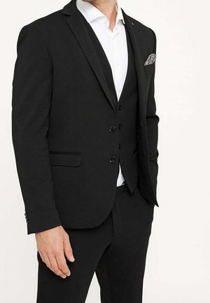 Hombre vestido con traje negro con chaleco, camisa blanca de vestir y un pañuelo con estampado en el bolsillo, de pie frente a un fondo claro y liso.