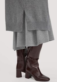 Graues, schichtweise gestricktes Kleid mit gerippten Bündchen und einem seitlichen Schlitz, kombiniert mit knielangen, dunkelbraunen, ledernen Blockabsatzstiefeln mit Reißverschluss.