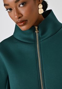 Veste teal à col montant avec une fermeture éclair dorée et une texture douce. Le mannequin porte des boucles d'oreilles rondes en or. Gros plan sur le vêtement et les accessoires.