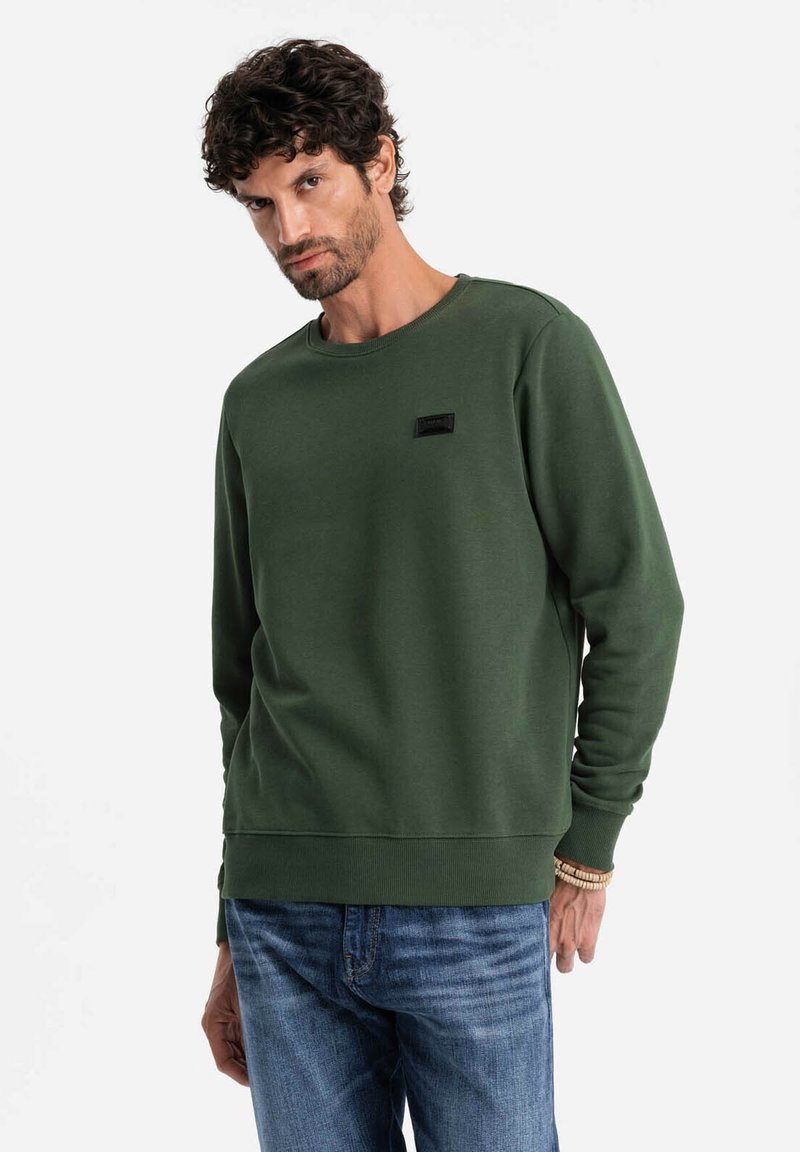 Felpa verde scuro con collo rotondo, polsini e orlo a coste, con un piccolo etichetta nera sul petto. Indossata con jeans blu.