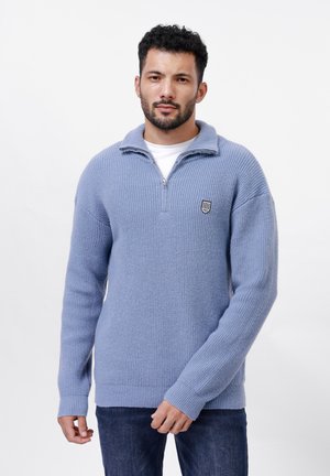 Suéter de punto azul claro con un cuello alto, media cremallera en la parte delantera, textura acanalada y un pequeño parche con logo en el lado izquierdo del pecho.