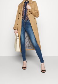 Beige Trenchcoat mit doppelreihigen Knöpfen, blumenmuster Bluse, schmale blaue Jeans und schwarze High-Heels. Hält eine beige Tasche.