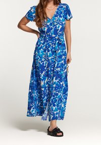 Donna che indossa un vestito sopra il ginocchio a fiori blu e bianchi con bottoni e vita legata, sandali neri senza chiusura, mentre tiene una tracolla a catena.
