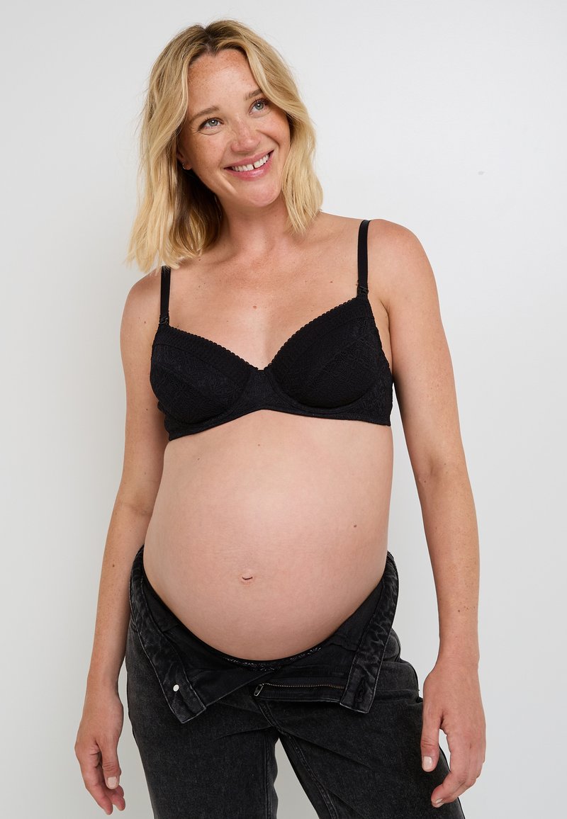 Reggiseno premaman nero con texture in pizzo e spalline regolabili. Il modello indossa jeans neri aperti, che mettono in evidenza un pancione evidente.