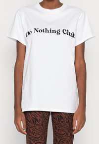 Vit bomullströja med en böjd svart textdesign "Do Nothing Club" över bröstet, med en avslappnad passform och korta ärmar.