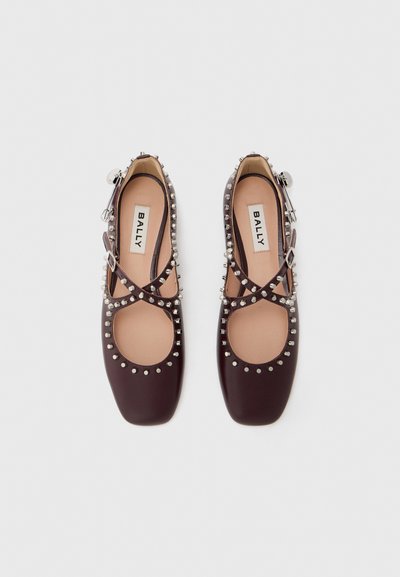 Ballerines en cuir bordeaux avec un bout rond, des sections découpées et des clous métalliques décoratifs le long des bords. Une sangle ajustable ajoute du détail.