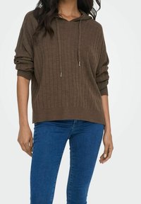 Kvinna iförd en brun ribbad tröja med dragband och blå jeans, stående mot en enkel vit bakgrund.