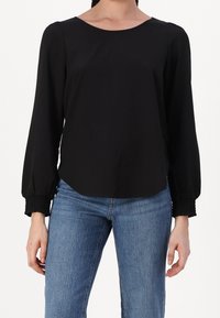 Blouse noire à manches longues avec un décolleté arrondi, des poignets élastiques et une texture de tissu lisse. Assortie à un jean bleu.