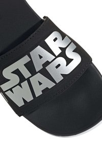 Svart slide-sandal med ett texturerat band som har den silverfärgade logotypen "STAR WARS", vilket kontrasterar mot en slät svart innersula.