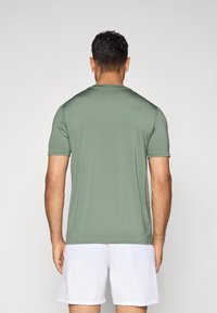 Homme avec des cheveux courts et foncés portant une chemise unie verte à manches courtes et un short blanc, se tenant de dos devant un fond uni.