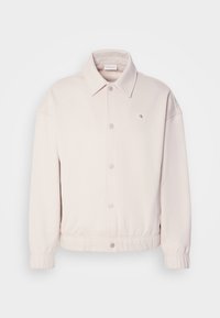 Giacca beige chiaro realizzata in tessuto morbido, con collo a camicia, bottoni a scatto, polsini elasticizzati e una piccola etichetta del logo sul lato sinistro del petto.