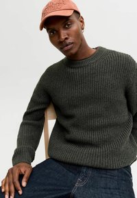Dark green ribbed knit sweater with a crew neckline, worn with blue denim jeans; model seated on a light wooden chair.