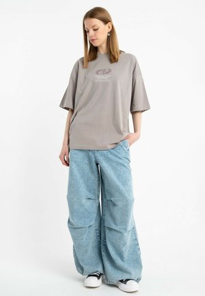 Młoda kobieta w oversize'owej szarej koszulce z napisem, szerokich jasno-niebieskich dżinsach i czarno-białych trampkach, stoi z jedną ręką w kieszeni.