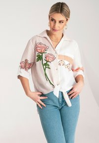 Pasiasty top w białym i jasnoróżowym kolorze z haftem kwiatowym, z guzikami z przodu, podwiniętymi rękawami i wiązanym przodem. Połączony z jasnoniebieskimi dżinsami.