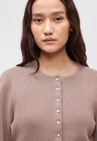 Ανοιχτό καφέ πλεκτό κάρντιγκ με στρογγυλή γραμμή λαιμού, που διαθέτει κλείσιμο με κουμπιά μπροστά και ριμπέ ύφανση στα μανίκια.